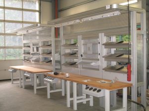 Montagearbeitsplätze aus der Industrie mit vier Tischen mit Holzplatte und Werkzeughalter am Tisch, hinter den Tischen hohe Regalsysteme aus Aluminiumprofilen mit schrägen Bauteilregalen und Beleuchtung für die Tische - Arbeitsplatzgestaltung, Layoutplanung, Steigerung der Produktivität eines Arbeitsplatzes